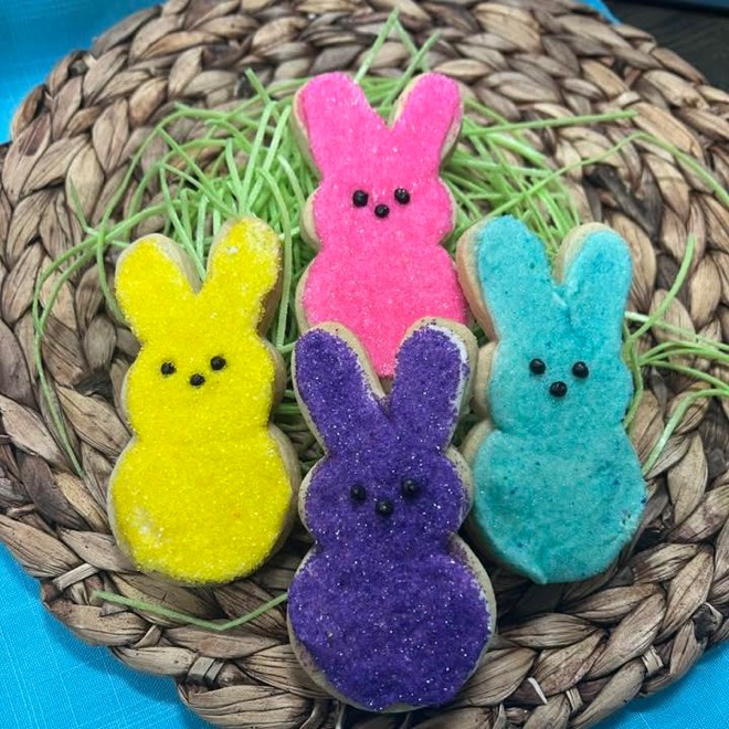 Three sugar-frosted bunny cookies in yellow, pink and teal, lined up on a woven mat with paper grass.
