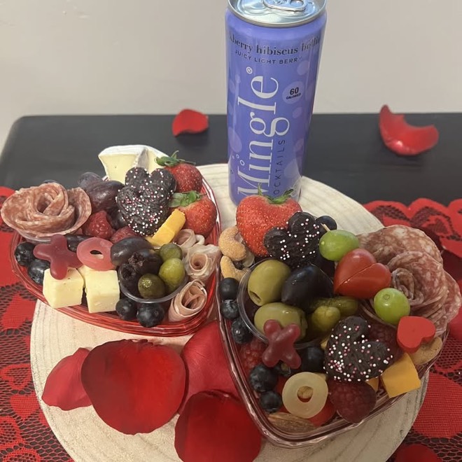 Two small heart-shaped charcuterie boats holding meat roses, cheese cubes, olives, berries and chocolate, beside a canned mocktail on a red runner.
