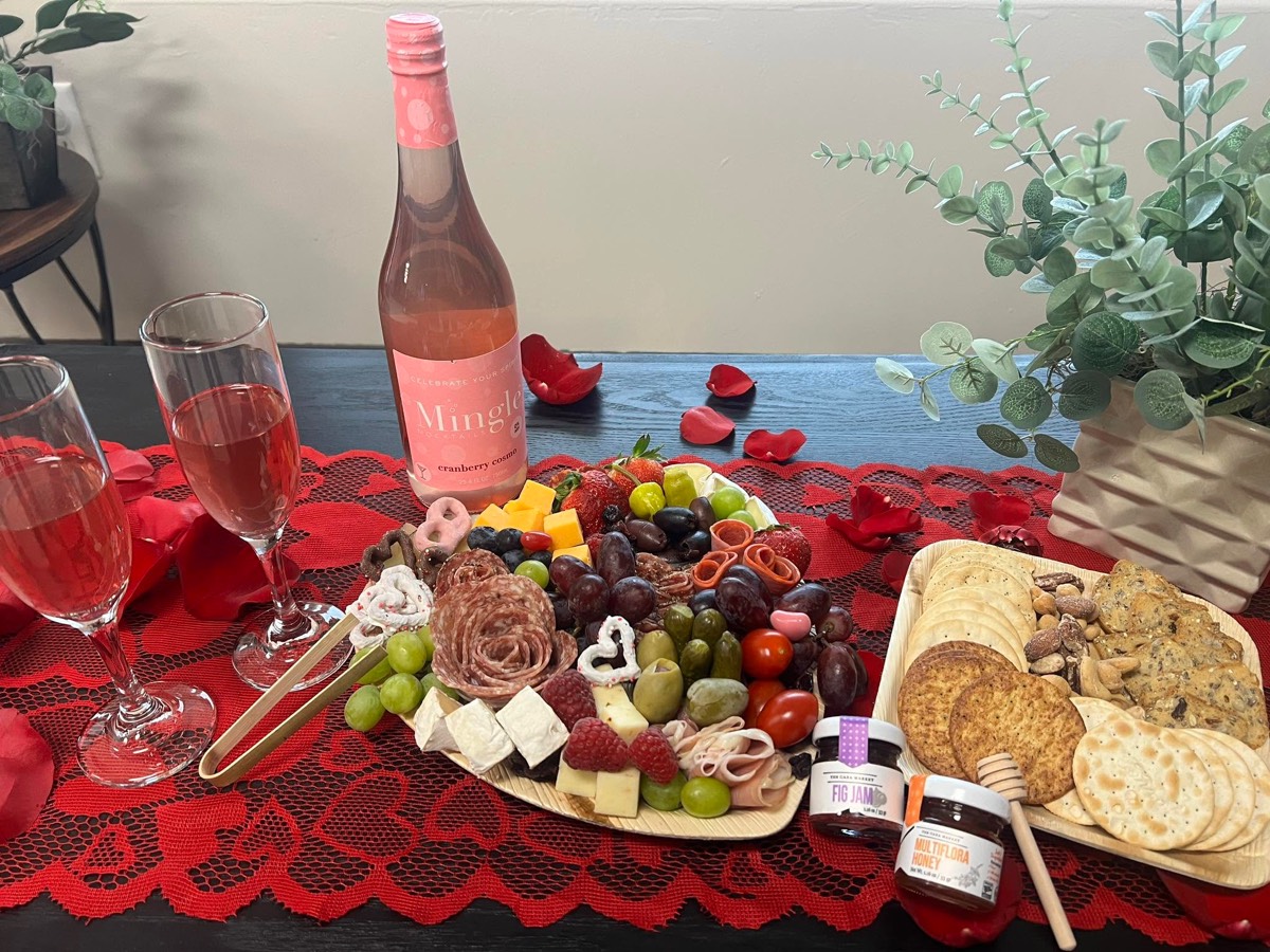 A Valentine's board on a red lace runner with a bottle of rose, two filled champagne flutes, meat roses, cheese cubes, grapes, crackers and chocolate-dipped treats, with eucalyptus behind.